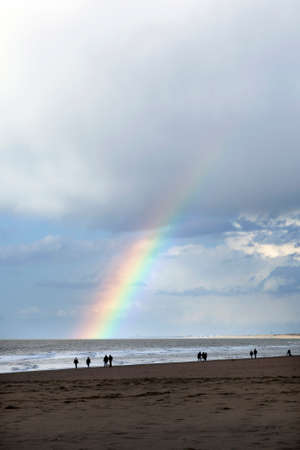 people stroll on north sea beach in holland near scheveningen with cloudy sky and rainbowの写真素材