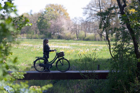 young blond girl with bicycle amongst fresh green spring foliage and meadow full of flowers in the netherlandsの写真素材