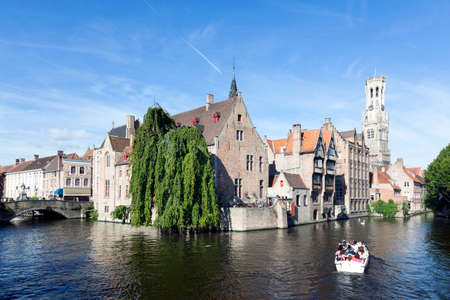 old houses and tourist boat in canal in centre of medieval Brugge in Belgium with blue sky in summerのeditorial素材