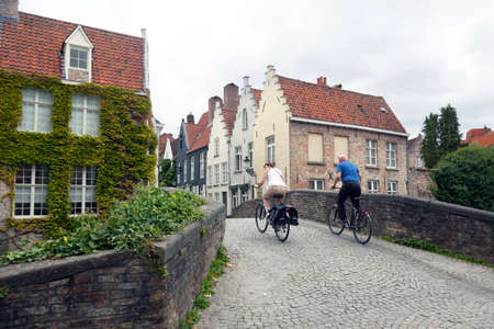 man and woman on bicycles try to ride over old medieval bridge in center of brugge in belgiumのeditorial素材