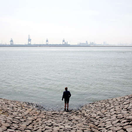 boy looks over Nieuwe Waterweg at harbour cranes near rotterdam harbourの写真素材