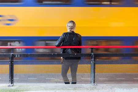 Young woman in black leather coat waits on platform of railway station in the netherlands while yellow and blue train passesの写真素材