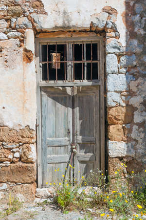very old wooden door with padlock in ancient stone wall of greek house on sunny day with yellow spring flowersの写真素材