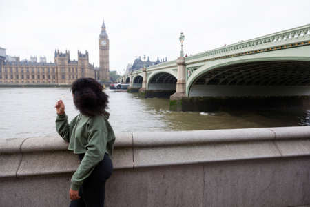 girl in green sweater leans on railing on thames embankment under westminster bridge and looks at houses of parliamentのeditorial素材