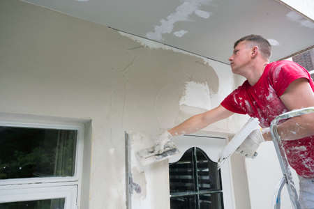 plasterer in red shirt works on white plaster of old house during insulation workの写真素材
