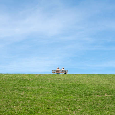 couple sit together on bench placed on top of grassy dike in friesland under blue sky