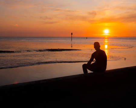 silhouette of sitting young man who looks over sea during sunset in dutch province of frieslandの写真素材