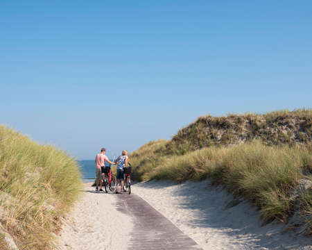 norderney, germany, 25 august 2019: couple with bicycles at top of dunes near sea on german island of norderney on sunny summer day with blue skyのeditorial素材