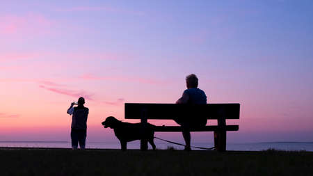 man takes pictures with moblie phone of colorful sunset while woman sits with dog on benchの写真素材