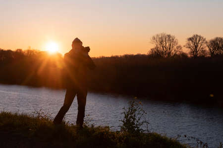 silhouette of man against the sun with camera at sunset in nature surroudings with canalの写真素材