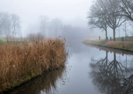 valleikanaal or valley canal and men on bicycle near scherpenzeel in the netherlands on misty winter dayの写真素材