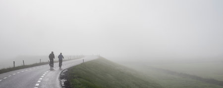 couple on bicycle rides in mist on dike near river rhine in hollandの写真素材