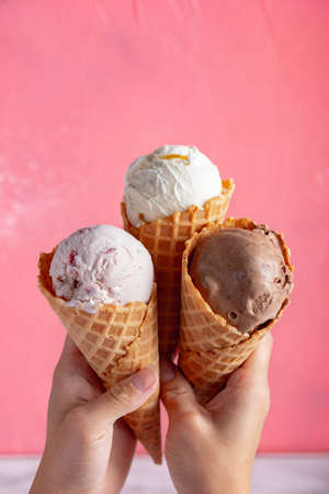 hands holding strawberry, vanilla, chocolate ice cream with waffle cone on pink backgroundsの写真素材