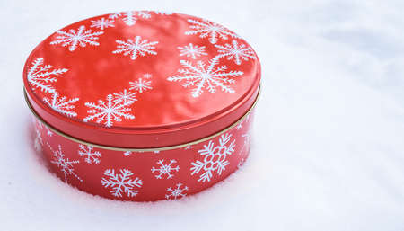 Red, round cookie & baked goods tin container decorated in red with white snowflake print pattern, sitting in natural snow.の写真素材