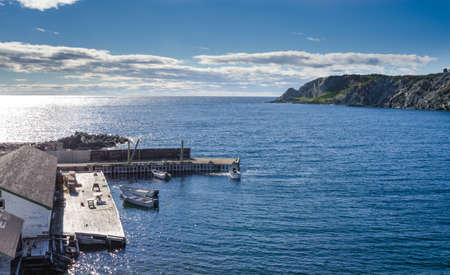 Twillingate, Newfoundland, man works on the engine of one of 4 boats tied up to a wooden, cliff-side dock house for the day, bright sunshine on calm coastal water.の写真素材