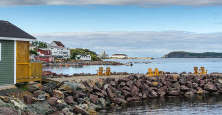 Golden adirondack chairs on a rock jetty.   Houses on the sea along a village shoreline.  Rural Newfoundland, Canada.の写真素材