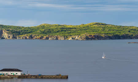 Late afternoon sun shines on an outcropping of high rugged terrain in calm sea water as a sailboat under motor power, goes along in the bay at Twillingate, Newfoundland, Canada.の写真素材