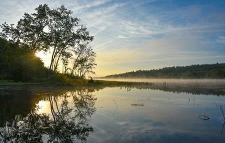 Brilliant and bright mid-summer sunrise on a lake.   Warm water and cooler air at daybreak creates misty fog patches.  Still water along a calm, quiet Ontario lakeside.の写真素材