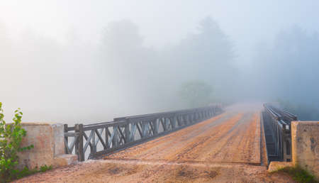 Low cloud in bright mist. surreal coloured subdued light over a single lane road.  Steel & timber bridge, spanning narrow lake water passage.の写真素材