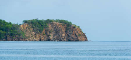 Trees and foliage grow straight out from very old rock on a steep, rising outcropping that borders a beach in Costa Rica.  Boat goes along rocky shores of Pacific coast.の写真素材