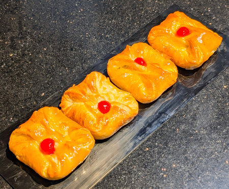 Four golden Danish pastries with glossy cherry topping arranged neatly on a black serving plate....