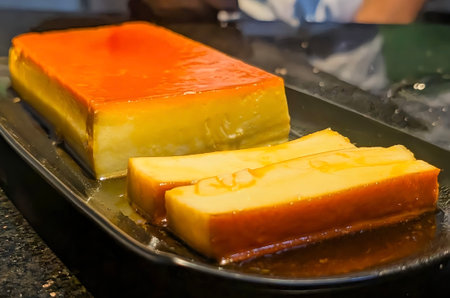 Close-up of a rich caramel flan dessert served on a black plate. The smooth custard and glossy...