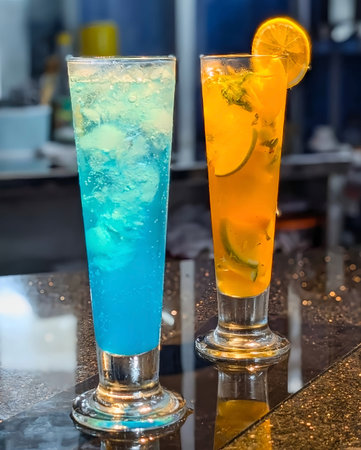 Two tall glasses of colorful summer mocktails â one bright blue lagoon and one orange citrus drink with lemon slices â served on a glossy table.の写真素材
