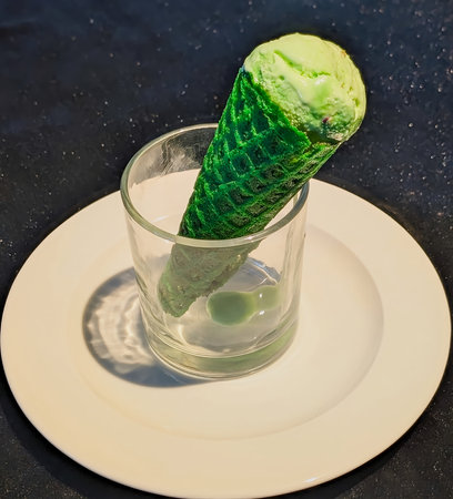 Creative dessert presentation featuring a vibrant green ice cream cone placed in a clear glass on a white plate over a dark background.の写真素材