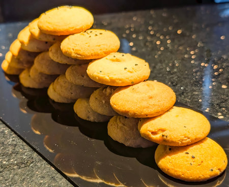 A vertical, close-up shot captures multiple stacks of freshly baked, golden-hued cookies, notably those topped with black sesame seeds, arranged diagonally on a highly reflective black platter.の写真素材