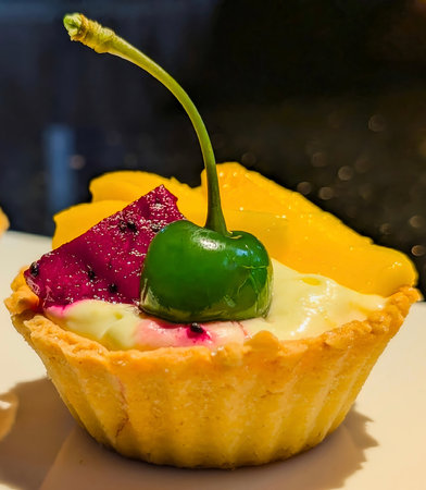 Vibrant mini fruit tarts featuring flaky shortcrust pastry filled with creamy custard, topped with fresh mango, purple dragon fruit, and a glossy green cherryの写真素材