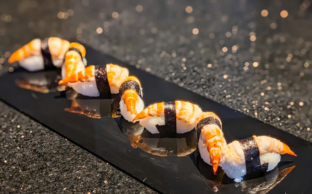 Five pieces of ebi nigiri sushi featuring cooked shrimp over vinegared rice, secured with seaweed strips and arranged on a glossy black rectangular plate.の写真素材