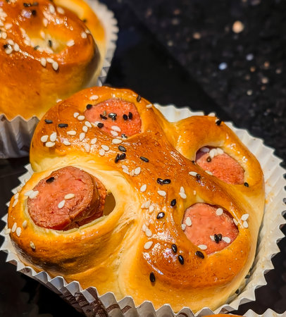 A close-up of a golden-brown sausage bun topped with white and black sesame seeds, served in a...