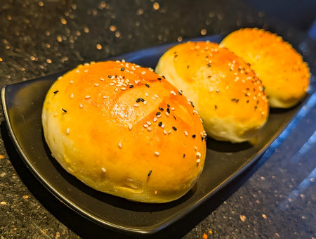 Three freshly baked bread rolls with glossy golden crusts topped with black and white sesame seeds.