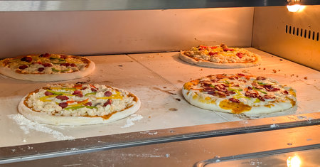 Four uncooked pizzas topped with sausage, bell peppers, and cheese bake directly on a floured stone...