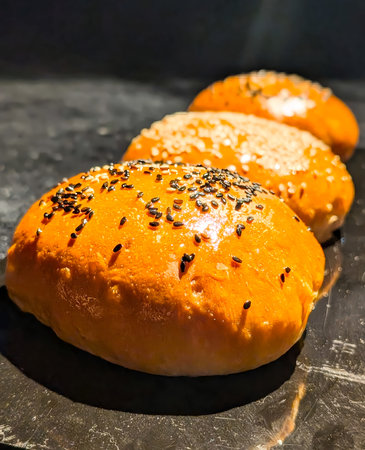Three golden-brown bread rolls with glossy crusts, topped with black and white sesame seeds,...