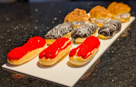 Selection of Ã©clairs topped with red frosting and chocolate glaze with white drizzle, alongside...