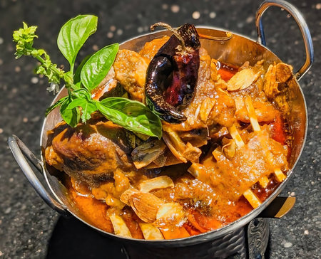 A richly spiced lamb curry served in a traditional metal bowl with handles, garnished with garlic,...