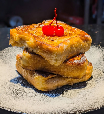 Three thick slices of golden-brown French toast stacked on a dark plate and dusted with fine...