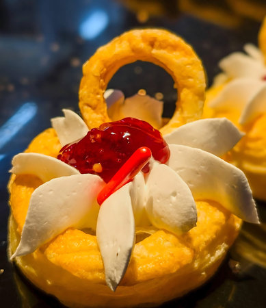 Glazed choux pastries filled with smooth yellow custard and topped with whipped cream and glossy red...