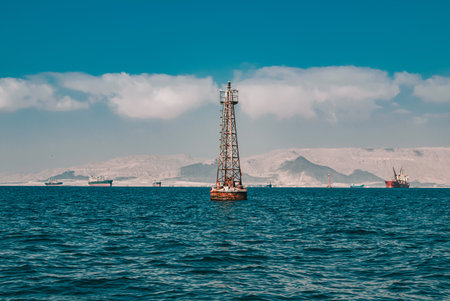 A wide seascape showing a lone navigational beacon standing in the blue sea with multiple cargo ships and a mountain range on the distant horizon under a cloudy skyの写真素材