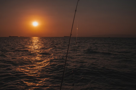 A solitary fishing rod is silhouetted against a beautiful warm sunset casting a golden reflection across the tranquil sea as distant ships sit on the horizonの写真素材