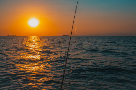 The sun sets in a blaze of orange and yellow over the sea creating a brilliant reflection on the water with a fishing rod in the foreground and ships on the horizonの写真素材
