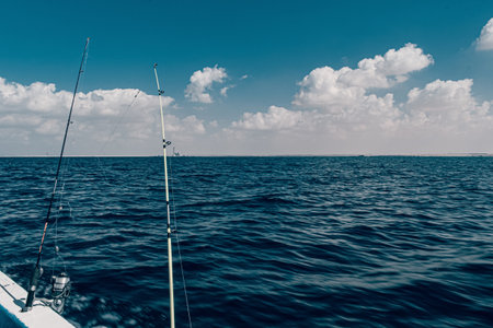 From the side of a boat two fishing rods are ready for action overlooking the vast deep blue waters of the sea under a beautiful sky with scattered white cloudsの写真素材