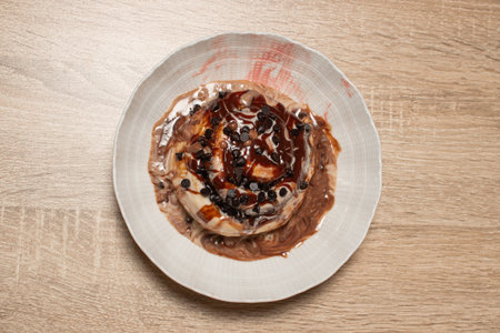 A top down flat lay photo of a single delicious cinnamon roll with chocolate sauce and chocolate chips in a white bowl on a light wood table with copy spaceの写真素材