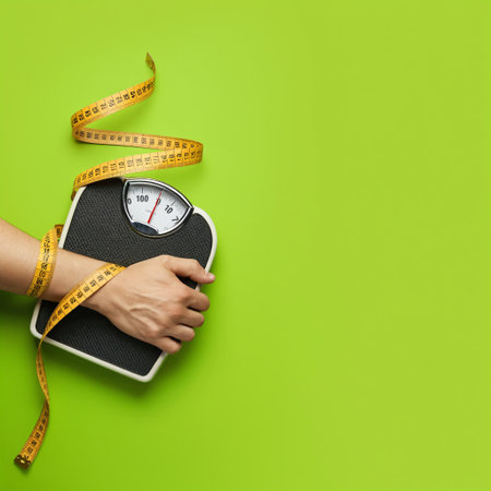 A person's hand holds a bathroom scale with a yellow measuring tape wrapped around their arm on a green background with copy space for health and diet contentの素材