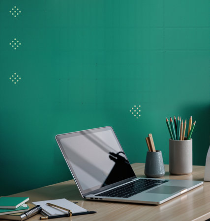 A clean and stylish home office or creative workspace featuring an open laptop on a wooden desk with notebooks and pens against a solid textured green wallの素材
