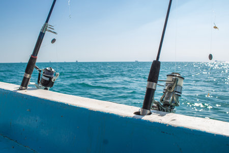 Close up of two fishing rods with spinning reels secured in holders on the side of a boat The sunny day reflects off the blue water of the Red Sea and distant cargo ships are on the horizonの写真素材