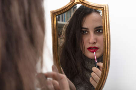Beautiful woman applying red lipstick using a handheld gold framed vintage mirror with focus to her reflection in the glassの写真素材