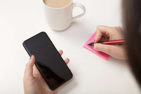 Woman making a sticky note reminder as she copies something off her mobile phone in an over the shoulder view to the blank screenの写真素材