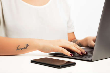 Hands of a young woman typing on a laptop computer in a close up view with a mobile phone alongside. With âla reineâ tattoo on arm, meaning â queenâ  in french  with crown signの写真素材